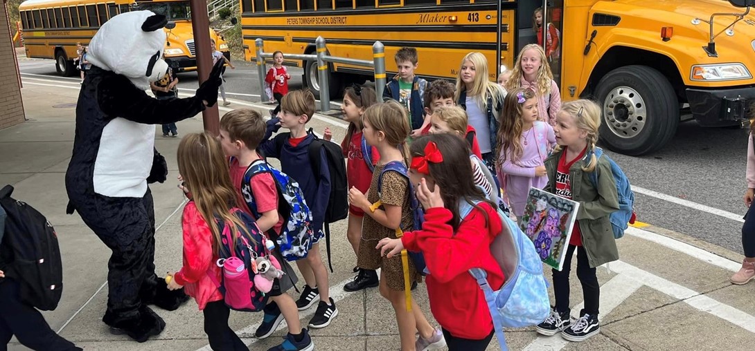Adult in panda costume gives a high-five to students getting off the bus